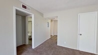 This single-story unit offers a practical and efficiently arranged floor plan accommodating three bedrooms and a kitchen within a compact footprint. Neutral beige walls and white trim are consistent throughout, creating a clean, cohesive aesthetic. The living area is carpeted and features a front entry door alongside a window fitted with horizontal blinds, allowing natural light to brighten the space.

The kitchen is well-appointed with white cabinetry offering ample upper and lower storage, gray laminate countertops, a double-basin white sink with a modern pull-down faucet, a white electric coil range, a full-size refrigerator, and a dishwasher. Wood-look vinyl flooring runs through the kitchen, adding durability in this high-use area.

The spacious primary bedroom features carpet flooring, neutral walls, and access to a bathroom. A walk-in closet with multiple built-in shelving tiers provides substantial storage capacity. The two additional bedrooms are carpeted and each include windows with blinds that admit natural light.

A hallway connects the bedrooms and living areas, finished with wood-look flooring for continuity. The textured ceilings, visible HVAC vents, and consistent trim work throughout reflect a straightforward, functional design suited to everyday living.