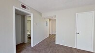 This single-story unit offers a practical and efficiently arranged floor plan accommodating three bedrooms and a kitchen within a compact footprint. Neutral beige walls and white trim are consistent throughout, creating a clean, cohesive aesthetic. The living area is carpeted and features a front entry door alongside a window fitted with horizontal blinds, allowing natural light to brighten the space.

The kitchen is well-appointed with white cabinetry offering ample upper and lower storage, gray laminate countertops, a double-basin white sink with a modern pull-down faucet, a white electric coil range, a full-size refrigerator, and a dishwasher. Wood-look vinyl flooring runs through the kitchen, adding durability in this high-use area.

The spacious primary bedroom features carpet flooring, neutral walls, and access to a bathroom. A walk-in closet with multiple built-in shelving tiers provides substantial storage capacity. The two additional bedrooms are carpeted and each include windows with blinds that admit natural light.

A hallway connects the bedrooms and living areas, finished with wood-look flooring for continuity. The textured ceilings, visible HVAC vents, and consistent trim work throughout reflect a straightforward, functional design suited to everyday living.