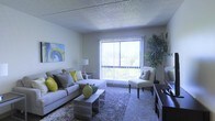 This single-floor space centers around a comfortable living room designed to maximize natural light and openness. The room features wall-to-wall carpeting in a neutral tone, complemented by a textured popcorn ceiling that adds a classic touch to the interior. A large sliding glass door dominates one wall, framed by vertical blinds that allow for adjustable light control while offering pleasant views of the surrounding greenery outside. The living area is well-proportioned, providing ample room for seating arrangements and furnishings. A sectional sofa with accent pillows in yellow and grey anchors the space, paired with a sleek glass and metal coffee table and an accent chair positioned near the window. A tall indoor plant adds a natural, organic element to one corner of the room, enhancing the overall ambiance. Wall-mounted artwork and a side table with a lamp contribute to the warm, livable atmosphere. A low-profile media console is partially visible along one side, suggesting a functional layout suited for everyday living. The overall aesthetic is casual and inviting, with a neutral palette accented by pops of color throughout.