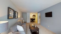 This single-story residence offers an open-concept layout that connects the living and dining areas in a practical, flowing arrangement. The living room is finished with plush wall-to-wall carpeting in a warm neutral tone, providing a comfortable foundation for the space. Soft gray walls create a calm, understated backdrop that complements the room's furnishings. A dark wood coffee table and coordinating gray sofa anchor the living area, while framed artwork and a wall-mounted flat-screen television add functional decorative touches to the walls.

The space opens directly into the dining area, which features warm-toned flooring and is illuminated by a ceiling fan with integrated lighting, providing both air circulation and ambient light. A dark wood high-top dining table with matching bar-style chairs serves as the focal point of this area. A mirror hung on the dining room wall enhances the sense of depth. A door visible in the background suggests access to a closet or utility area. The overall ambiance is clean, neutral, and well-organized, making the space feel welcoming and easy to personalize.