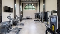 This fitness center is a striking, single-floor facility defined by its soaring ceilings and industrial-inspired architectural design. Tall, dark steel columns line the perimeter, creating a bold contrast against the white brick walls and lending the space a refined, contemporary aesthetic. A grand arched window at one end of the room frames lush outdoor greenery, bathing the workout floor in abundant natural light, while additional upper-level windows further enhance the bright, open atmosphere.

The facility is well-equipped to support a comprehensive workout routine. A row of treadmills and elliptical machines runs along one side of the room, ideal for cardiovascular training. On the opposite side, a multi-station cable machine and various strength-training apparatus provide ample options for resistance work. A dedicated free weights area features a fully stocked dumbbell rack alongside an adjustable bench, set on a protective rubber mat. Flat workout benches are also distributed throughout the space for versatility.

Wall-mounted flat-screen televisions are positioned at eye level to keep exercisers engaged during their sessions. The polished concrete flooring is both durable and visually clean, complementing the overall modern industrial character of the space. A staircase visible toward the rear suggests access to an upper-level mezzanine, adding further dimension to this well-appointed fitness facility.
