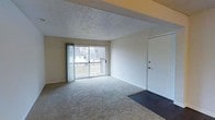 This single-floor residence offers a clean and functional layout designed for comfortable everyday living. The main living area is finished with wall-to-wall carpeting in a neutral tone, providing a warm and inviting atmosphere. A textured ceiling adds classic character to the space, while wide sliding glass doors serve as the focal point of the room, flooding the interior with natural light and providing direct access to a private balcony. The balcony overlooks a surrounding outdoor area, offering pleasant views of the exterior greenery and neighboring rooftops. Vertical blinds frame the sliding doors, allowing for adjustable light control and privacy. The entry door, positioned to the side of the living area, features a peephole and is finished in a clean white, consistent with the room's neutral color palette. The walls are painted in a simple off-white tone that enhances the brightness of the space and creates a versatile backdrop. Overall, the space presents a straightforward, well-lit interior with practical finishes and a seamless connection to the outdoors through its balcony access.