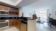 This single-floor space showcases a thoughtfully designed open-concept layout that seamlessly connects the kitchen and living area. The kitchen is appointed with sleek black granite countertops that extend across a peninsula, providing both workspace and a natural divider between the cooking and living zones. Warm, medium-toned wood cabinetry lines the kitchen walls, offering ample storage, while built-in appliances including a dishwasher and cooktop contribute to a streamlined aesthetic. Light tile flooring runs throughout, lending a clean, cohesive feel to the space. A recessed ceiling detail above the kitchen adds architectural interest. The adjoining living area benefits from floor-to-ceiling windows that flood the space with natural light and frame views of lush greenery and an outdoor balcony, creating a bright and airy ambiance. Decorative touches such as a table lamp, potted plants, and a woven basket add warmth. The overall design balances functional layout with an open, welcoming atmosphere suitable for both everyday living and entertaining.