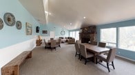 Located at 126 W Berryhill Dr in Grass Valley, California, this single-story space offers an open, airy layout defined by soaring vaulted ceilings and soft aqua-blue walls accented by white beadboard wainscoting throughout. The main gathering area is expansive and versatile, seamlessly blending multiple functional zones into one cohesive environment. A striking floor-to-ceiling stone fireplace serves as the architectural centerpiece, anchoring the lounge area where a large sectional sofa and round coffee table create an inviting seating arrangement bathed in natural light from a bank of windows. Adjacent to the lounge, a large rustic wood dining table with upholstered chairs accommodates generous group gatherings, while a secondary dining or activity table with additional seating provides further flexibility. A wall-mounted flat-screen TV and a wooden media console with decorative marquee lighting add modern functionality to the space. The kitchen features warm honey-toned wood cabinetry, light countertops, a window overlooking the outdoors, and includes appliances such as a range, dishwasher, and microwave. Neutral carpet flooring flows throughout the main areas, while the kitchen transitions to light hardwood-style flooring. Recessed lighting throughout ensures a bright, welcoming ambiance suitable for a variety of gatherings or events.