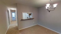 This single-floor space is designed with a practical and functional layout that maximizes connectivity between living areas. The main living space features neutral beige carpeting underfoot and smooth gray walls complemented by crisp white baseboards, creating a clean and understated aesthetic. A textured ceiling adds subtle visual interest overhead, while a wall-mounted multi-bulb light fixture provides focused illumination.

A standout architectural feature is the pass-through window cut directly into the wall between the living area and kitchen. Framed in white with a dark wood ledge serving as a small bar or serving counter, this opening allows light and sightlines to flow between spaces while maintaining a sense of definition between rooms. Through the pass-through, the kitchen is visible and features a white refrigerator, white range with overhead ventilation hood, and wooden cabinetry.

A hallway extends from the living area, with a door at the far end suggesting additional rooms beyond. The overall ambiance is modest and neutral, offering a blank canvas well-suited for customization and comfortable everyday living.