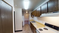 This single-floor unit at 1500 S Adams Ave in Marshfield, Wisconsin, offers a straightforward, functional layout well-suited for everyday living. The galley-style kitchen runs along one wall and is equipped with dark wood cabinetry providing ample upper and lower storage, light wood laminate countertops, a double-basin sink, a coil-burner electric range, and a full-size refrigerator. Tile-style flooring runs through the kitchen and hallway, creating a clean transition between spaces.

The living area is generously sized, featuring neutral carpet, a ceiling fan with light fixture, and a large vertical-blind window that allows natural light and outdoor views of surrounding greenery. Baseboard heating units are present throughout the unit, including in the living room and bedrooms. The bedrooms mirror each other in design — both carpeted, with vertical-blind windows overlooking outdoor scenery and baseboard heaters beneath each window.

The bathroom includes a single-sink vanity with wood cabinetry, a lighted mirror, an over-toilet storage cabinet, and a shower-tub combination with a curtain. Dark wood interior doors are consistent throughout, complementing the kitchen cabinetry. The overall aesthetic is clean and practical, with a neutral color palette across all rooms.