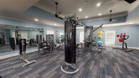 This single-floor fitness center is thoughtfully designed to support a comprehensive workout experience across multiple dedicated activity zones. The space is finished with soft blue walls, crisp white crown molding, and white baseboards, lending a clean and polished aesthetic throughout. Durable patterned carpet tiles in blue-gray tones cover the floors, providing both comfort and style underfoot.

The strength training area is generously appointed with a Matrix functional trainer as its centerpiece, complemented by a cable crossover system, selectorized weight machines, a multi-function rack with bench, and an assortment of stability balls and resistance accessories. Large wall-mounted mirrors amplify the sense of space and allow for proper form monitoring during workouts. Ceiling fans with recessed lighting ensure comfortable air circulation and consistent illumination.

A separate cardio zone along the windowed perimeter houses treadmills and an elliptical trainer, benefiting from natural light and views to the outdoors. A wall-mounted flat-screen television is positioned nearby for entertainment during cardio sessions, and a water station provides convenient hydration access.

The cycling studio features multiple indoor cycling bikes arranged in an open area, with its own ceiling fan and direct access through double exit doors. Throughout the facility, the layout is open and uncluttered, offering ample space to move freely between equipment stations.