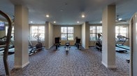 Located at 13250 Westheimer Rd in Houston, Texas, this fitness center is part of the Emilo community and is designed to offer residents a comprehensive workout experience on a single level. The space is thoughtfully laid out, with distinct zones accommodating a wide variety of exercise needs. A robust selection of strength-training equipment includes cable machines, weight benches, and a multi-station pull-up and dip tower, while a well-stocked dumbbell rack provides a full range of free weights for targeted training. The cardio section features treadmills, elliptical trainers, and rowing machines, each positioned to take advantage of the natural light streaming in through large windows that offer views of the surrounding community. Wall-to-wall mirrors amplify the sense of space and allow users to monitor their form throughout their workout. Ceiling fans throughout the room contribute to a comfortable environment, while recessed lighting ensures the space is well-illuminated. Mounted flat-screen televisions are positioned throughout the fitness center for entertainment during workouts. The flooring consists of durable, patterned carpet suited for a high-traffic fitness environment. Entry to the center is through a glass-paneled door, accompanied by posted fitness center policies from Emilo, ensuring a well-managed and welcoming facility for all residents.
