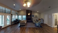 Located at 1255 W Pleasant Run Rd in Lancaster, Texas, this single-story community space offers an open, welcoming layout with a range of functional areas designed for both work and relaxation. The main living area is anchored by soaring vaulted ceilings and a dramatic floor-to-ceiling stone fireplace, creating a striking focal point. Rich hardwood-style flooring flows throughout, adding warmth and visual continuity across the space. Natural light pours in through an abundance of windows fitted with horizontal blinds, and a set of French doors opens directly to an outdoor pool area, seamlessly connecting the interior to the exterior. The entry features a classic arched wood door with an oval glass inset, setting an inviting tone upon arrival. A casual seating arrangement with a sofa and patterned accent chairs centers the living area around a decorative area rug. Adjacent to this, a dining nook offers a round table with upholstered chairs, ideal for small gatherings. The space also includes a dedicated workstation area with desks and computer setups, as well as a separate private office room for more focused work. A small bar-height counter and kitchenette area are visible near the entry, adding convenience. Ceiling fans with light fixtures provide additional illumination throughout this versatile, multi-purpose community space.