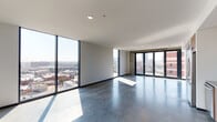 This contemporary high-rise unit offers a striking industrial-modern aesthetic defined by polished concrete floors, exposed concrete accent walls, and an open single-floor layout that maximizes both space and natural light. Expansive floor-to-ceiling windows wrap around the main living areas, framing panoramic views of the surrounding urban skyline and flooding the interior with abundant daylight throughout the day.

The open living area is spacious and versatile, anchored by the dramatic window walls and complemented by large sliding metal panels that add a bold, architectural character to the space. A set of glass doors provides direct access to an outdoor balcony area, further enhancing the connection to the cityscape beyond.

The kitchen is thoughtfully designed with warm wood cabinetry, stainless steel appliances including a range, dishwasher, and refrigerator, paired with a grey tile backsplash and sleek countertops that balance the industrial rawness of the surrounding concrete elements.

The bathroom presents a refined, spa-like atmosphere featuring a glass-enclosed shower, dark geometric tile walls, a wall-mounted sink with a metal-framed vanity, and open wood shelving that adds warmth and practical storage. Throughout the unit, the interplay of raw concrete, warm wood tones, and abundant natural light creates a sophisticated and distinctly urban living environment.
