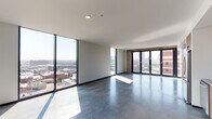 This contemporary high-rise unit offers a striking industrial-modern aesthetic defined by polished concrete floors, exposed concrete accent walls, and an open single-floor layout that maximizes both space and natural light. Expansive floor-to-ceiling windows wrap around the main living areas, framing panoramic views of the surrounding urban skyline and flooding the interior with abundant daylight throughout the day.

The open living area is spacious and versatile, anchored by the dramatic window walls and complemented by large sliding metal panels that add a bold, architectural character to the space. A set of glass doors provides direct access to an outdoor balcony area, further enhancing the connection to the cityscape beyond.

The kitchen is thoughtfully designed with warm wood cabinetry, stainless steel appliances including a range, dishwasher, and refrigerator, paired with a grey tile backsplash and sleek countertops that balance the industrial rawness of the surrounding concrete elements.

The bathroom presents a refined, spa-like atmosphere featuring a glass-enclosed shower, dark geometric tile walls, a wall-mounted sink with a metal-framed vanity, and open wood shelving that adds warmth and practical storage. Throughout the unit, the interplay of raw concrete, warm wood tones, and abundant natural light creates a sophisticated and distinctly urban living environment.