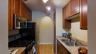 This single-floor residence offers a thoughtfully designed layout centered around a galley-style kitchen that serves as a functional and visually appealing heart of the home. The kitchen is outfitted with warm, rich wood cabinetry that lines both sides of the space, providing generous storage throughout. Granite-style countertops offer ample prep space, complemented by a stainless steel double sink and a full suite of appliances, including a gas range and a stainless steel refrigerator. Track lighting runs along the ceiling, casting bright, even illumination throughout the kitchen. A standout architectural feature is the elegant arched passageway at the far end of the kitchen, which leads seamlessly into the adjoining living areas. This arched detail adds a classic, timeless character to the home's interior design. Light hardwood flooring runs throughout, enhancing the warm and inviting ambiance of the space. The clean, neutral wall tones provide a versatile backdrop that complements the rich wood tones of the cabinetry. Overall, this single-floor home combines practical functionality with charming architectural details, making it a comfortable and characterful living space.