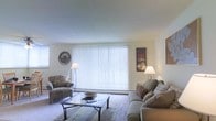 This single-floor residence offers a clean, comfortable living environment designed with functionality and ease in mind. The living room serves as the centerpiece of the space, featuring a large picture window dressed with vertical blinds that allow for adjustable natural light, brightening the room with a soft, diffused glow. The walls are finished in a crisp, neutral white, creating a versatile backdrop that enhances the sense of openness throughout. Plush wall-to-wall carpeting in a warm neutral tone adds comfort underfoot and contributes to the room's relaxed ambiance. The living area is well-appointed, with seating that includes a cushioned sofa accented with decorative pillows and a coordinating patterned armchair, arranged around a sleek glass-top coffee table. Tasteful artwork adorns the walls, adding visual interest and a personal touch to the space. Floor lamps provide warm supplemental lighting, creating a cozy atmosphere during evening hours. The overall design aesthetic is approachable and unpretentious, emphasizing comfort and livability. With its bright, airy feel and thoughtfully arranged furnishings, this residence provides a welcoming retreat suited to everyday living.