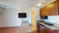 This single-floor studio offers a thoughtfully arranged layout that maximizes every square foot of available space. The main living area is anchored by rich, warm-toned hardwood flooring that extends throughout, creating a cohesive and inviting atmosphere. A ceiling fan with an integrated light fixture provides both comfort and illumination, while a large window dressed with vertical blinds allows natural light to fill the room. A wall-mounted flat-screen TV and a window-unit air conditioner round out the living space's functional features.

The kitchenette is efficiently designed and tucked neatly along one wall, featuring warm wood cabinetry, granite countertops, a stainless steel sink, a compact refrigerator, a microwave, and a portable cooktop. A breakfast bar-style granite counter peninsula serves as both a workspace and casual dining surface, seamlessly dividing the kitchen from the living area. Upper cabinetry provides ample storage overhead.

A closet space is accessible just off the main area, offering additional storage. The bathroom is clean and simply appointed, with white tile walls, a bathtub and shower combination with a curtain rod, and a toilet — presenting a functional, well-maintained space. The unit is accessed through a solid, dark-painted entry door with a modern keypad lock.
