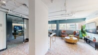 This contemporary office suite offers a well-appointed single-floor layout that balances open collaboration with private workspace. The design aesthetic blends industrial and modern elements, most notably through the exposed white-painted ceiling infrastructure — including visible ductwork, conduit, and track lighting — which lends an urban, loft-like character to the space.

The open lounge area serves as the heart of the suite, featuring rich teal accent walls, large windows that invite abundant natural light and views of surrounding greenery, and a distinctive geometric-patterned carpet in warm orange and neutral tones that anchors the space visually. Mid-century modern seating, including cognac leather armchairs and a round wood coffee table, contribute to a refined yet welcoming atmosphere.

A glass-walled private office provides transparency and openness while maintaining acoustic separation. It is equipped with a dedicated workstation and display area, making it well-suited for focused work or client consultations. Sliding glass doors and full-height glass partitions further reinforce the open, airy feel throughout.

Structural columns are integrated cleanly into the layout, and warm accent lighting complements the natural light from the window line. The overall ambiance is professional yet inviting, combining functional design with stylish, curated details that reflect a modern, design-forward workplace.