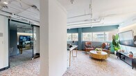 This contemporary office suite offers a well-appointed single-floor layout that balances open collaboration with private workspace. The design aesthetic blends industrial and modern elements, most notably through the exposed white-painted ceiling infrastructure — including visible ductwork, conduit, and track lighting — which lends an urban, loft-like character to the space.

The open lounge area serves as the heart of the suite, featuring rich teal accent walls, large windows that invite abundant natural light and views of surrounding greenery, and a distinctive geometric-patterned carpet in warm orange and neutral tones that anchors the space visually. Mid-century modern seating, including cognac leather armchairs and a round wood coffee table, contribute to a refined yet welcoming atmosphere.

A glass-walled private office provides transparency and openness while maintaining acoustic separation. It is equipped with a dedicated workstation and display area, making it well-suited for focused work or client consultations. Sliding glass doors and full-height glass partitions further reinforce the open, airy feel throughout.

Structural columns are integrated cleanly into the layout, and warm accent lighting complements the natural light from the window line. The overall ambiance is professional yet inviting, combining functional design with stylish, curated details that reflect a modern, design-forward workplace.