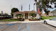 This single-story rental office is a well-maintained, standalone building designed to serve as a professional leasing and management hub. The exterior features a classic pitched roofline with warm-toned stucco walls and textured finish, lending the structure a clean and approachable appearance. The building's façade is framed by an array of tall, mature palm trees and lush, colorful flowering plants — including vivid reds, pinks, and yellows — that line the walkway leading to the front entrance, creating an inviting and well-kept first impression. Decorative banner flags flank the entry path, adding a sense of energy and visibility. The front entrance is fitted with a glass-paneled French-style door that allows natural light to filter into the interior, with informational signage and brochure holders mounted near the entry for visitor convenience. The building is ADA-compliant, with designated accessible parking spaces directly in front. An exterior drop box is mounted on the stucco wall beside the entrance, providing a practical after-hours solution for document submissions. The surrounding parking area is paved and well-marked, with additional greenery and palm trees enhancing the outdoor environment. The overall ambiance is professional, organized, and welcoming — well-suited for a residential community office setting.