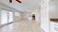 This two-story space is designed with an open, airy layout that seamlessly connects the living area and kitchen on the main floor. Light-toned ceramic tile flooring runs throughout, reflecting natural light and contributing to the overall bright, clean aesthetic. The expansive living area features a textured ceiling with a ceiling fan, track lighting, and multiple windows fitted with blinds. A set of French doors opens to the exterior, flooding the space with additional natural light and providing convenient outdoor access.

The kitchen is positioned at the heart of the main floor and features white cabinetry, granite-toned countertops, and a full suite of stainless steel appliances, including a range, microwave, and refrigerator. A peninsula island offers additional counter space and visual separation from the living area. Nearby, a dedicated closet-style laundry area houses a full-size washer and dryer. A bathroom on the main level includes white cabinetry with stone-finish countertops and a large mirror. A staircase leads to the second floor, expanding the overall living space across two levels.