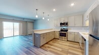 This single-floor unit offers a thoughtfully designed open-concept layout that seamlessly connects the kitchen and living area. The kitchen is a standout feature, outfitted with shaker-style cabinetry in a soft off-white finish, light quartz countertops, and a full suite of stainless steel appliances including a range, over-the-range microwave, dishwasher, and refrigerator. A peninsula with pendant lighting overhead creates a natural divide between the kitchen and the expansive living area, which is finished with durable wood-look plank flooring in a cool gray-blue tone. The living space receives natural light and provides access to a private balcony through sliding glass doors, offering outdoor views. Bedroom areas feature plush carpeting and bright windows with blinds. The unit includes two bathrooms, each finished with gray shaker-style vanities, white quartz countertops, wood-look tile flooring, and well-lit mirrors. One bathroom features a walk-in shower, while the other includes a shower-tub combination. Neutral wall tones and recessed lighting throughout contribute to a clean, modern ambiance.