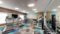 This single-floor fitness center is thoughtfully designed to support a wide range of workout routines. The space is anchored by a bold, floor-to-ceiling photographic wall mural depicting a sunlit coastal pier, creating an energizing and visually expansive atmosphere. Complementing the mural, the walls are painted in a refreshing aqua blue, accented by wood-paneled sections that add warmth and texture to the overall aesthetic.

The flooring features a striking geometric hexagonal carpet pattern in shades of teal, blue, and gray, adding a modern, dynamic feel underfoot. Overhead, multiple flush-mount circular light fixtures provide bright, even illumination throughout the space.

The cardio zone is well-stocked with treadmills, elliptical trainers, and cycling bikes, while a flat-screen television is mounted on the wall for entertainment during workouts. A dedicated strength training area includes a multi-function cable machine, a full dumbbell rack, and a weight bench. Additional accessories such as medicine balls, resistance bands, exercise mats, and stability balls are neatly organized on wall-mounted racks.

Full-length mirrors line one wall, enhancing the sense of space and allowing users to monitor their form. A window provides natural light, and the layout allows for comfortable movement between equipment zones throughout the room.