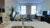 This thoughtfully designed single-floor space showcases a seamlessly integrated open-plan layout that maximizes both functionality and comfort. The kitchen features a polished granite countertop with a sleek stainless steel sink and modern faucet, flowing naturally into the dining and living areas beyond.

The dining area is furnished with a dark wood table and matching chairs, anchored by a stylish zebra-print area rug that adds a touch of visual interest. The living space beyond is warm and inviting, centered around a cushioned sofa and armchair with a low coffee table. Lush indoor plants add a refreshing natural element to the interior.

Two generously sized windows fitted with horizontal blinds line the far wall, allowing ample natural light to fill the space while offering views of the surrounding building exterior. Light-toned hardwood flooring runs throughout, lending a clean, cohesive aesthetic across the entire floor plan.

A dedicated workspace is thoughtfully positioned near the windows, complete with a desk and ergonomic chair, making this space well-suited for both relaxing and productive activities. The neutral color palette of soft greens and whites creates a calm, airy ambiance throughout.