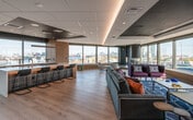 This striking single-floor space is designed with a high-end, contemporary aesthetic that seamlessly blends functionality with biophilic design. Large, floor-to-ceiling windows wrap around multiple areas of the floor, flooding the interiors with natural light and offering sweeping panoramic views of the surrounding cityscape and waterfront.

The open-plan lounge area features light hardwood flooring, recessed and cove lighting with warm accent tones, and a carefully curated selection of colorful, modern furnishings arranged across multiple seating zones. A built-in window bench runs along the perimeter, inviting occupants to take in the city views.

A fully equipped kitchen area includes a substantial island with bar seating, warm wood cabinetry, and professional-grade appliances — designed to support both casual gatherings and catered events.

One of the most distinctive features is a dedicated indoor garden atrium enclosed by glass partitions. This lush sanctuary houses tall tropical plants, living green walls, and a dramatic deep-blue decorative water feature, all set beneath a dark sculptural ceiling — creating a tranquil, nature-inspired retreat within the urban setting.

An additional lounge nook features a raised terrazzo platform, a sleek built-in fireplace framed in dark stone, and intimate seating arranged to capture the city panorama. The space also includes a media wall and reception-style service counter, rounding out a versatile, world-class environment.