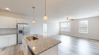 This single-floor residence offers a well-designed open-concept layout with light-toned luxury vinyl plank flooring flowing throughout the main living areas. The kitchen serves as the focal point of the space, showcasing a central island with granite countertops and a built-in sink, illuminated by elegant gold pendant lights. White shaker-style cabinetry lines the walls, complemented by a classic white subway tile backsplash and a full suite of stainless steel appliances, including a refrigerator, electric range with over-the-range microwave. The kitchen opens seamlessly into the living area, which features a ceiling fan and a window that brings in natural light. The primary bedroom is generously proportioned, with plush carpeting, multiple windows, a ceiling fan, and sliding door access to an outdoor space. It connects to a private bathroom with wood-look flooring and a walk-in closet equipped with wire shelving. A secondary bathroom features a white vanity with a quartz countertop, a large mirror, and brushed nickel fixtures. A carpeted walk-in closet with wire rack shelving provides additional storage. Recessed lighting and a neutral color palette throughout contribute to a clean, contemporary ambiance.