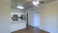 This single-floor residence showcases an open-concept layout that seamlessly connects the kitchen and living areas, creating a spacious and functional environment. The kitchen is outfitted with stainless steel appliances, including a refrigerator, electric range, and microwave, all complemented by white cabinetry and light granite-style countertops. A peninsula with a built-in sink provides additional workspace and serves as a natural divider between the kitchen and the living area. Crown molding runs along the ceiling throughout the space, adding a refined architectural detail, while a ceiling fan with a light fixture provides both air circulation and ambient lighting. The flooring features dark hardwood-style planks that extend across the main living area, offering a warm and contemporary aesthetic. A doorway visible from the main area leads to an adjacent room with carpeted flooring, suggesting a comfortable private space. The neutral wall tones and clean white trim contribute to a bright, cohesive atmosphere throughout the unit.