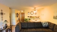 This single-floor residence offers a comfortable and functional open-concept layout that seamlessly connects the living, dining, and kitchen areas. The living room serves as the heart of the home, anchored by a large, plush sectional sofa and accented with decorative touches including framed wall art and an oval mirror. Lush houseplants add a vibrant, natural element throughout the space. A ceiling fan with integrated lighting is centrally positioned overhead, providing both comfort and warm illumination across the open area. The dining area features a round white table with matching chairs, creating a welcoming spot for meals. The kitchen is visible beyond, equipped with countertop appliances and a tiled backsplash, designed for everyday practicality. A hallway with a wooden door leads to additional areas of the home, adding depth to the floor plan. The walls are painted in warm tones that contribute to the cozy ambiance, and wall-to-wall carpeting runs throughout the living areas, adding softness underfoot. Decorative accents, including a large wall clock and script wall art, personalize the space and reflect a homely, comfortable style throughout.