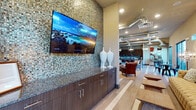This single-floor communal space is thoughtfully designed to blend contemporary and industrial aesthetics across several interconnected areas. The layout flows seamlessly from a media and buffet zone into a central lounge and onward to a dining or bar area, creating a versatile environment suited for both relaxed gatherings and social events.

The media wall serves as a striking focal point, clad floor-to-ceiling in multi-toned mosaic glass tile in earthy greens, golds, and browns. A large flat-screen television is mounted above dark espresso cabinetry finished with granite countertops, offering functional storage alongside decorative white ceramic vases.

The central lounge area features elevated ceilings with exposed silver ductwork lending an industrial character, complemented by globe pendant lighting and recessed fixtures. A generously sized tufted linen sofa anchors the seating arrangement alongside rich leather accent chairs, all positioned over wood-look striped flooring. The accent wall displays a bold collection of circular mixed-media art pieces, while clerestory windows admit natural light. A bistro nook with floor-to-ceiling windows provides outdoor views. Restroom facilities are accessible via a well-appointed corridor with coordinating wood-tone tile flooring and dark-framed glass doors.