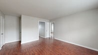 This single-story space is defined by a consistent design language carried throughout every room. Rich, dark hardwood-style flooring runs continuously across all areas, providing warmth and visual cohesion against the neutral gray walls and crisp white trim and baseboards.

The living area is open and generously proportioned, with a window allowing natural light to filter in and illuminate the space. The kitchen is well-equipped, featuring white shaker-style cabinetry extending to the upper walls, granite-look countertops, a double basin sink, and white appliances including a refrigerator and electric range with an overhead vent hood. The layout provides ample storage and counter workspace.

Multiple bedrooms are present throughout the unit, each outfitted with ceiling fans, closet storage — including sliding and louvered door options — and windows that bring in natural light and outdoor views. The bedrooms maintain the same neutral palette and hardwood-style flooring found elsewhere in the space.

The bathroom includes a full tub-shower combination with tiled surround, a toilet, and a window for ventilation and light. The overall ambiance of the space is clean, functional, and move-in ready, with a cohesive aesthetic maintained from room to room.