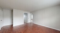 This single-story space is defined by a consistent design language carried throughout every room. Rich, dark hardwood-style flooring runs continuously across all areas, providing warmth and visual cohesion against the neutral gray walls and crisp white trim and baseboards.

The living area is open and generously proportioned, with a window allowing natural light to filter in and illuminate the space. The kitchen is well-equipped, featuring white shaker-style cabinetry extending to the upper walls, granite-look countertops, a double basin sink, and white appliances including a refrigerator and electric range with an overhead vent hood. The layout provides ample storage and counter workspace.

Multiple bedrooms are present throughout the unit, each outfitted with ceiling fans, closet storage — including sliding and louvered door options — and windows that bring in natural light and outdoor views. The bedrooms maintain the same neutral palette and hardwood-style flooring found elsewhere in the space.

The bathroom includes a full tub-shower combination with tiled surround, a toilet, and a window for ventilation and light. The overall ambiance of the space is clean, functional, and move-in ready, with a cohesive aesthetic maintained from room to room.
