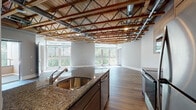 Located in Watertown, MA, this single-floor unit offers a compelling blend of industrial character and modern finishes. The defining architectural feature is the soaring exposed wood truss ceiling, which spans the entire space and gives it a loft-like, open feel. Visible ductwork and steel piping complement the raw aesthetic, while recessed lighting ensures the interiors remain warm and well-lit.

The open-concept living and kitchen area is anchored by a large central island with granite countertops and an undermount stainless steel sink. The kitchen is equipped with contemporary flat-front cabinetry in a warm wood tone, stainless steel appliances including a range, microwave, refrigerator, and dishwasher. Wide-plank hardwood flooring flows throughout the main living areas, while large floor-to-ceiling windows flood the space with natural light and offer views of the surrounding greenery.

The bedrooms continue the industrial design language with the same exposed truss ceilings and hardwood floors, and generous windows. The bathrooms feature tile flooring, granite-topped vanities, and full tub-shower combinations with clean, neutral finishes. The overall ambiance is modern and airy, with a distinctive character that sets this space apart.