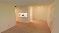 This single-floor space features an open and functional layout highlighted by neutral cream-toned walls and warm beige carpeting that extends throughout the living and adjoining areas. The kitchen is positioned behind a half-wall pass-through, which serves as both a visual divider and a casual dining or serving counter. The kitchen itself is bright, with white cabinetry, a white electric stovetop, and under-cabinet lighting that illuminates the workspace. A HVAC vent is visible in the ceiling, indicating a central air system. Beyond the main living area, an open doorway leads to a secondary room or hallway, suggesting additional functional space within the unit. The flush-mount ceiling light provides overhead illumination for the main living zone. The overall design is straightforward and practical, with clean lines and a minimal aesthetic. The open sight lines between the kitchen and living area contribute to a sense of connected, usable space, making the layout well-suited for everyday living and simple entertaining.