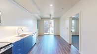 This efficiently designed one-bedroom, one-bathroom unit offers a thoughtful layout across a single floor, making excellent use of its compact footprint. The open-concept living and kitchen area is anchored by a striking kitchen featuring bold cobalt blue lower cabinetry paired with crisp white upper cabinets, white countertops, and a full suite of stainless steel appliances including a refrigerator, range, microwave, and dishwasher. Hardwood-style flooring runs throughout the main living area, enhancing the contemporary aesthetic and contributing to a bright, clean atmosphere. A ceiling fan is centrally positioned in the living space, which opens directly to a private balcony through a sliding glass door, inviting natural light and outdoor views into the unit. The bedroom is a comfortable, carpeted room with its own ceiling fan and a window that further extends the sense of airiness. Neutral cream-toned walls throughout provide a versatile backdrop. The bathroom includes a full soaking tub and shower combination, a vanity with a white countertop, and blue cabinetry that ties back to the kitchen's design palette, creating a cohesive visual identity across the unit. Overall, the space presents a modern and functional living environment with consistent design details throughout.
