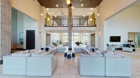 Located at 3110 Grand Ave in Pinellas Park, Florida, this contemporary community clubhouse is a striking amenity space defined by its dramatic double-height ceilings and open, airy floor plan. Light-toned large-format tile flooring runs throughout, reflecting the abundant natural light that pours in through floor-to-ceiling windows and glass sliding doors that frame views of the surrounding tropical landscaping and palm trees.

The expansive main lounge area serves as the heart of the space, furnished with multiple oversized sectional sofas arranged around low coffee tables, creating inviting conversation areas beneath a cascading arrangement of globe pendant lights. A mezzanine level with sleek metal railings overlooks the main floor, adding architectural depth and visual interest.

A full-service kitchen features a generous island with bar stool seating, stainless steel appliances, white cabinetry, and playful pendant lighting. Adjacent bar and lounge zones offer additional counter seating with a mounted television, exposed ductwork, and a relaxed industrial-modern aesthetic. A dramatic marble-clad accent wall anchors a linear fireplace flanked by additional lounge seating. A sun-drenched dining area with high bistro tables sits beside glass walls, completing this well-appointed and versatile gathering space.