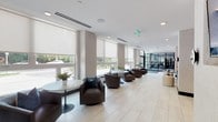 The lobby at 1077 Warburg Ave in Yonkers, NY presents a polished, contemporary design that sets a welcoming tone for the building. Light-toned large-format tile flooring extends throughout the space, contributing to a clean, open aesthetic. A row of oversized windows fitted with roller shades lines one side of the lobby, flooding the interior with natural light while offering views of the surrounding greenery outside.

The seating areas are thoughtfully arranged to encourage both casual conversation and quiet relaxation. Dark brown swivel chairs are paired with round marble-top pedestal tables along the windowed wall, while a vibrant navy blue sofa adorned with colorful accent pillows anchors a lounge area on the opposite side, complemented by cylindrical cream-colored ottomans. A bold abstract canvas painting mounted against a warm wood-paneled wall adds an artistic focal point to this seating nook.

Textured light gray wall panels provide visual depth throughout the lobby, while recessed lighting ensures consistent illumination. Elevator banks finished in metallic tones are conveniently accessible near the lounge area. At the far end of the lobby, a glass-enclosed fitness center with steel-framed partitions is clearly visible, housing cardio equipment and offering a seamless connection between the lobby and building amenities.
