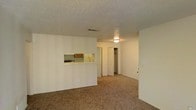 This single-story space in Midland, Texas, presents an open and functional layout designed for comfortable everyday living. The main living area is finished with wall-to-wall carpet in a neutral tan tone, lending warmth to the room while contributing to a quiet, understated atmosphere. Textured ceilings run throughout, adding a classic architectural detail consistent with the overall design.

A half-wall pass-through connects the kitchen to the living space, offering a sense of openness while maintaining a defined cooking area. The kitchen features wooden cabinetry and a gas range, visible through the pass-through opening. White lower cabinetry and a light countertop complete the kitchen's clean, practical aesthetic.

Toward the back of the main living area, a door opens to reveal a generously sized closet with interior rod and shelving space, providing ample storage. A flush-mount ceiling light fixture illuminates the space, supplemented by a centrally placed HVAC vent for climate control. The neutral wall tones and simple finishes throughout create a clean, move-in ready environment well-suited to a variety of furnishing styles.