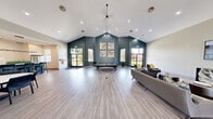 This well-appointed single-floor community space offers a generous and open floor plan designed for both socializing and relaxation. The interior is defined by dramatic vaulted ceilings that rise to impressive heights, filling the main gathering area with an airy, expansive atmosphere. Light-toned wood-look flooring runs throughout, complementing the neutral white and bold teal accent walls that give the space a modern, contemporary character.

The main lounge area features a full-size pool table positioned beneath a striking teal accent wall adorned with decorative artwork. Large windows on multiple sides invite an abundance of natural light and provide views of the surrounding greenery. A comfortable seating area with a sofa, accent chairs, and patterned rugs anchors one side of the room, while a wall-mounted flat-screen TV adds an entertainment focal point.

A dedicated entryway with glass double doors leads into the space, flanked by a smaller sitting nook with leather chairs and sculptural décor. The fully equipped kitchen showcases a large central island with bar seating, white quartz countertops, a subway tile backsplash, stainless steel appliances, and a modern statement chandelier. Adjacent to the kitchen, a clearly marked coffee bar provides a convenient amenity. Decorative wall-mounted disc art adds artistic detail throughout the space.