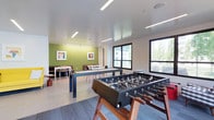 This single-floor recreation space is thoughtfully designed to serve as a social and entertainment hub. The room features a clean, modern aesthetic anchored by large windows that flood the interior with natural light and offer views of the surrounding exterior. Recessed lighting and sleek linear ceiling fixtures provide ample illumination throughout.

The space is divided into distinct activity zones. A full-size foosball table takes center stage near the windows, while an air hockey table provides additional gaming options in the middle of the room. The flooring transitions between large-format tile and patterned carpet, helping to define the various areas.

A dining or lounge area is positioned toward the back of the room, furnished with a long white table and seating, set against a bold green accent wall adorned with framed artwork. A cozy booth-style seating nook is tucked into the corner, adding an intimate gathering spot.

The overall ambiance is energetic yet inviting, with a contemporary color palette of white, grey, and green creating a fresh and lively atmosphere. This well-appointed space is ideal for socializing, casual gatherings, and recreational activities.