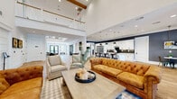 This impressive clubhouse-style gathering space blends transitional and modern farmhouse design elements into a cohesive, welcoming environment. The grand entry foyer sets the tone immediately, featuring warm hardwood-style flooring, a striking multi-tiered wrought iron chandelier, and a sweeping staircase with white balusters and a wood handrail leading to an open upper-level mezzanine. Soaring double-height ceilings with exposed wood beam accents amplify the sense of openness throughout the main living area.

The expansive great room is anchored by a tufted caramel leather Chesterfield sofa and complementary neutral seating, arranged around a rustic wood coffee table. A stone fireplace serves as a focal point at the far end of the room, adding warmth and character. The space flows seamlessly into a full gourmet kitchen outfitted with white shaker cabinetry, a dark herringbone tile backsplash, stainless steel appliances including a large built-in refrigerator, white quartz countertops, and a central island with bar seating beneath elegant pendant lights.

Adjacent café-style nooks with bistro tables and metal chairs, accented by bold slate-blue walls and curated abstract artwork, provide intimate gathering spots. Recessed lighting and large glass entry doors allow natural light to fill the interior, creating a bright and inviting atmosphere throughout.