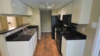 This single-floor space showcases a well-organized galley-style kitchen as its central feature. The kitchen is designed with functionality in mind, featuring a symmetrical layout with white cabinetry lining both sides, providing ample upper and lower storage. Dark granite-look countertops offer a striking contrast against the white cabinets, creating a bold visual aesthetic throughout the space.

On one side, a double stainless steel sink is set into the countertop, accompanied by additional lower cabinet storage and a dishwasher. The opposite side hosts a full suite of black appliances, including an electric range with coil burners, a built-in microwave, and a full-size refrigerator, keeping all cooking essentials within easy reach.

Warm-toned hardwood flooring runs the length of the kitchen, adding a natural element that complements the overall color palette. Track lighting mounted on the ceiling provides direct illumination throughout the galley corridor. The textured ceiling adds dimension overhead, while neutral-toned walls contribute to a clean, cohesive look. The layout maximizes the use of space efficiently, creating a practical and visually cohesive cooking environment.