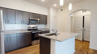 This single-floor residence offers a thoughtfully designed layout with contemporary finishes throughout. The kitchen is the centerpiece of the home, showcasing rich dark-espresso cabinetry that extends to the ceiling, providing generous storage space. Speckled granite countertops run along the full perimeter and continue onto a prominent center island, which features an undermount sink with a chrome faucet and serves as both a prep area and casual dining space. A white subway tile backsplash adds a clean, modern contrast to the dark cabinetry, while a full suite of stainless steel appliances — including a refrigerator, range, and built-in microwave — completes the kitchen. Warm hardwood-style flooring flows through the kitchen and main living areas, complementing the bright, neutral wall tones. Elevated ceilings and elegant pendant lighting over the island lend an airy, open feel to the kitchen, which connects to adjacent spaces through a graceful arched entryway. The bathroom mirrors the kitchen's design language, featuring matching granite countertops, dark cabinetry, an undermount sink, and a large framed mirror. The bedroom is a calm retreat, finished with plush wall-to-wall carpeting, a ceiling fan, and a window that invites natural light into the space.