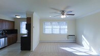 This single-floor space offers a clean, functional layout with a welcoming open-concept design. The living area is finished with wall-to-wall carpeting and features a ceiling fan with integrated lighting, recessed lighting, and crown molding that adds a polished touch to the room. A large window fitted with horizontal blinds allows an abundance of natural light to fill the space, creating a bright and airy atmosphere. A wall-mounted HVAC unit and a baseboard heating system provide year-round climate control. A decorative column elegantly separates the living area from the adjacent kitchen, which features tile flooring and dark cabinetry. The kitchen is equipped with a black refrigerator and appears to offer views to the outdoors through its own window. The neutral, light-toned walls throughout the space provide a versatile backdrop suitable for a variety of interior styles. The overall ambiance is practical and comfortable, with thoughtful details that contribute to a well-maintained and livable environment.