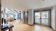 This thoughtfully designed single-floor clubhouse offers a refined and inviting atmosphere from the moment you step through its glass-paneled entrance. Light wood-style flooring flows throughout the entire space, providing a warm, cohesive foundation that ties each area together seamlessly.

The main lounge area commands attention with soaring vaulted ceilings accented by exposed dark wood beams, creating a striking architectural focal point. Large windows dressed in teal-trimmed frames invite an abundance of natural light and offer views of the surrounding outdoor areas. A generously sized sectional sofa anchors the lounge, complemented by accent chairs and flat-screen televisions mounted on a sleek, wood-paneled feature wall — ideal for relaxed gathering or casual viewing.

A dedicated social and dining area extends further into the space, featuring a long bar-height communal table with seating, banquette-style booth seating, wall-mounted displays, and warm pendant lighting. This versatile zone is well-suited for both social events and individual use.

The coffee bar area is a standout feature, showcasing a rustic reclaimed wood accent wall fitted with open shelving, a mirrored backsplash, and a countertop service station complete with coffee and beverage equipment. A live-edge wood communal table adds an organic, artisanal touch to this inviting corner of the space.
