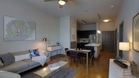 This single-floor space showcases a thoughtfully designed open-concept layout that seamlessly integrates the living, dining, and kitchen areas into one cohesive environment. Warm hardwood flooring flows throughout, anchoring the space with a sense of continuity and warmth.

The living area is bright and inviting, furnished with a comfortable sofa accented with decorative pillows and a glass-topped coffee table resting on a area rug. A ceiling fan with integrated lighting overhead provides both airflow and ambient illumination, while a sleek floor lamp adds a warm, layered glow to the corner.

Adjacent to the living room, the dining area features a wooden table with upholstered chairs, offering a practical yet stylish spot for meals. The dining space flows naturally into the kitchen, which is anchored by a peninsula counter with a clean white surface — ideal for casual seating or meal prep. Dark cabinetry lines the kitchen walls, creating a bold contrast against the lighter countertops and stainless steel appliances, including a full-size refrigerator and microwave. Track lighting and pendant fixtures illuminate the kitchen with precision and style.

A console table with decorative accessories adds a finishing touch near the living area, while neutral gray walls and modern design choices throughout give this space a polished, contemporary ambiance.