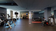 This fitness center offers a comprehensive workout environment designed to accommodate a wide range of exercise routines. The space is laid out across a single open floor, finished with durable rubber flooring that provides both traction and impact absorption throughout the workout areas.

The cardio zone features treadmills, elliptical machines, and rowing machines, complemented by a wall-mounted flat-screen television. Large windows along one side of the room flood the area with natural light and offer views of the surrounding exterior, creating an energizing atmosphere for cardio training.

The strength training section is equally well-appointed, with a full dumbbell rack, adjustable weight benches, and a cable machine system. A dedicated functional training area includes a rack of medicine balls, stability balls in various sizes, resistance bands, and open floor space for stretching and mat exercises — marked by a red yoga mat.

Full-length mirrors line one wall, enhancing the sense of space and allowing users to monitor their form. Linear LED lighting runs across the ceiling, providing bright, even illumination throughout. The color palette of deep blue-gray walls paired with neutral accents gives the space a clean, modern aesthetic. Overall, this fitness center is thoughtfully designed to support a full spectrum of fitness activities in a professional and motivating setting.