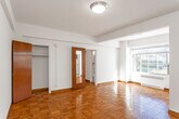 This single-floor residence offers a thoughtfully laid-out living space defined by warm, honey-toned parquet flooring that extends seamlessly throughout every room, lending a timeless character to the interior. The open living and dining area is bathed in natural light streaming through large windows that frame views of the surrounding cityscape, with a radiator unit positioned beneath the sill for year-round comfort. An additional window-mounted air conditioning unit ensures climate control during warmer months. Rich, warm-toned wood doors and trim provide a consistent design thread throughout the space, complementing the neutral white walls and clean architectural lines. Multiple closets with built-in rods offer ample storage, while structural beam details add visual interest to the ceilings. The kitchen, accessible directly from the main living area, features functional cabinetry and appliances. The bathroom is a standout feature, showcasing classic vintage tilework in a black-and-cream color palette — complete with a mosaic-patterned floor, bold navy tile accents, a mirrored medicine cabinet, a built-in bathtub with overhead shower, and a window for natural ventilation. The entry area provides a welcoming transition into the home, with convenient closet access immediately upon arrival.