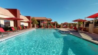 This single-story amenity space is designed for outdoor recreation and relaxed social gatherings, showcasing a well-appointed pool area complemented by a range of leisure features. The centerpiece is a generously sized swimming pool with crystal-clear turquoise water, accompanied by a separate circular spa/hot tub finished with blue mosaic tile detailing. A shallow wading ledge connects the two water features, adding a resort-like quality to the setting.

The surrounding pool deck is outfitted with red lounge chairs and coordinating red market umbrellas, providing ample shaded seating throughout the day. Shade sails extend over a dedicated lounge and dining zone, further enhancing comfort in the open-air environment. A built-in outdoor kitchen station features dual stainless steel grills with storage below, ideal for entertaining.

An interior lounge area, accessible from the pool deck, is enclosed with a striking stacked-stone accent wall in warm neutral tones. Skylights in the ceiling draw in natural light, brightening the space throughout the day. Red-and-white striped cushioned seating arranged around low coffee tables creates a comfortable gathering space. The architecture reflects a Southwestern-inspired style, characterized by terracotta roof tiles and warm earth-toned exteriors.