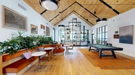 Located at 6000 Morris Pl in East Hanover, NJ, this generously sized clubroom offers an impressive single-floor layout designed for both relaxation and social engagement. The space is immediately defined by its dramatic vaulted ceilings clad in warm natural wood planking, accented by exposed black structural beams and pendant lighting that creates a refined yet inviting atmosphere throughout.

The open floor plan flows seamlessly across multiple functional zones. A central lounge area anchors the space with plush seating, including a large sectional sofa and leather chairs, arranged around a fireplace and wall-mounted television. Adjacent to this is a dedicated game area featuring a full-size billiards table set atop a bold area rug. A curved banquette seating area, surrounded by lush indoor plants and curated artwork, provides an intimate gathering spot ideal for casual meetings or conversation.

The kitchen bar area is appointed with dark cabinetry, bar seating, and pendant lighting, while black-framed glass partitions define a separate conference or private room. Light oak hardwood flooring runs throughout, complementing the white walls and wood ceiling. Floor-to-ceiling black-framed windows and glass doors flood the interior with natural light and offer views of the surrounding exterior. Abundant greenery throughout adds a vibrant, organic touch to this thoughtfully designed community amenity space.